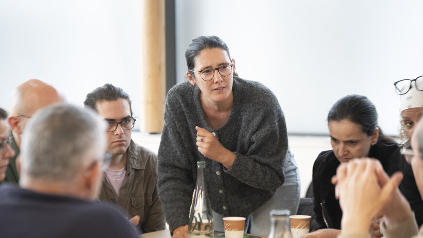 Gros plan sur une des tables. Une jeune femme s'est levée et parle à ses convives. 