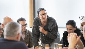 Gros plan sur une des tables. Une jeune femme s'est levée et parle à ses convives. 