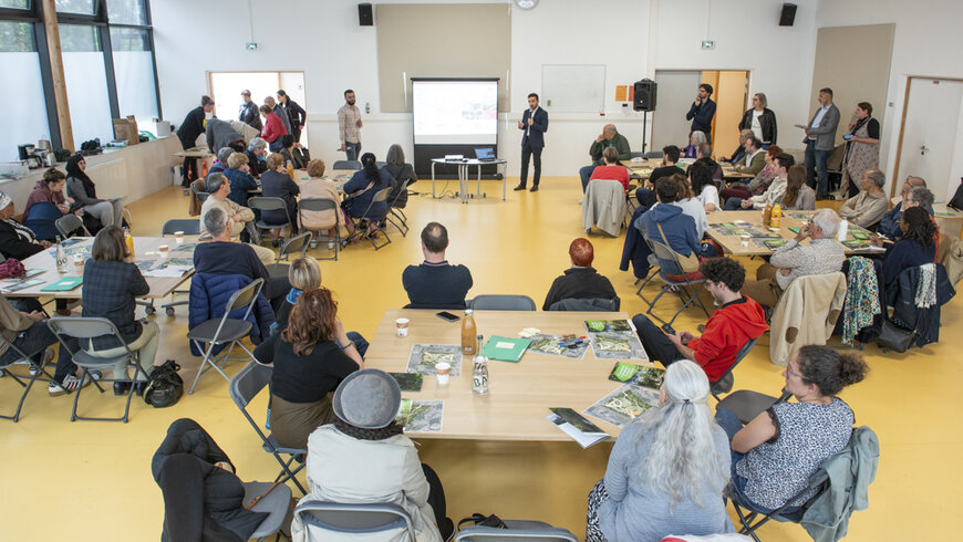 Une assemblée est réunie dans le préau de l'école Niki-de-Saint-Phalle. Ils sont installés autour de plusieurs tables. Dans le fond, un homme se tient debout devant un écran.
