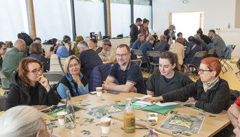 Gros plan sur une des tables. les gens regardent le plan du parc ainsi que des vignettes représentant des végétaux et différents élément de mobilier urbain. 