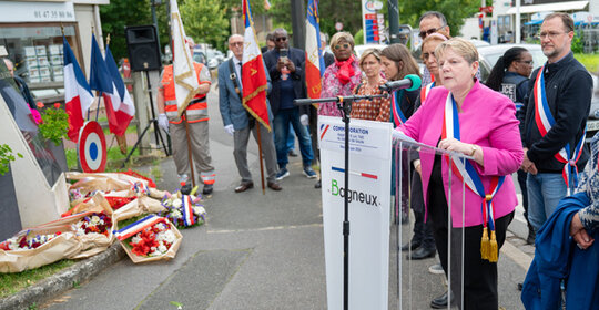 Marie-hélène Amiable devant un pupitre lit son discours lors d'une cmmémoration. Des gerbes de fleurs sont déposées au pied d'une stèle commémorative en granit. Dans le public des représentants des associations d'anciens combattants portent des drapeaux bleu balnc rouge..  